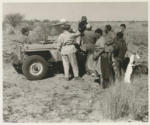 Group of people standing around the Jeep, including !Ungka wearing a dress, "Old Gau" wearing a shirt, and expedition members