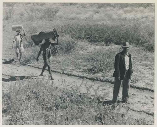 Two men walking with Wilhelm Camm and carrying his belongings