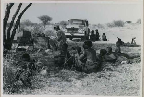 People sitting and reclining near Nicholas England at recording equipment and Chevrolet, long shot
