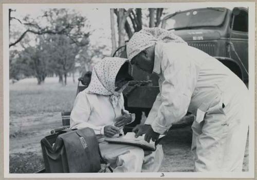 Lorna Marshall and Laurence Marshall wearing clothing and head coverings to protect against tsetse flies, and looking at Lorna's notebook

