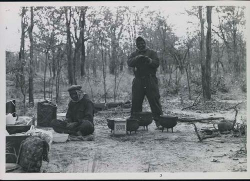 John Nambahu and Manuel sitting and standing in road kitchen and wearing clothing and head coverings to protect against tsetse flies
