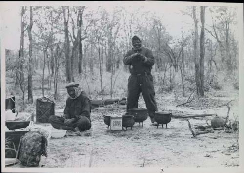 John Nambahu and Manuel sitting and standing in road kitchen and wearing clothing and head coverings to protect against tsetse flies
