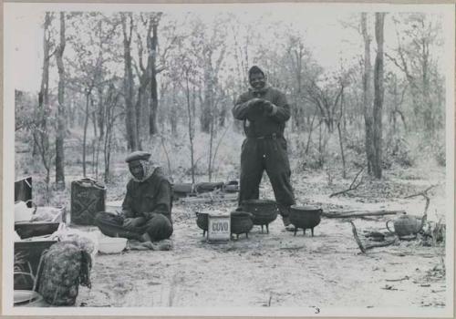 John Nambahu and Manuel sitting and standing in road kitchen and wearing clothing and head coverings to protect against tsetse flies
