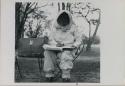 Lorna Marshall wearing clothing and head covering to protect against tsetse flies, sitting and writing in her notebook