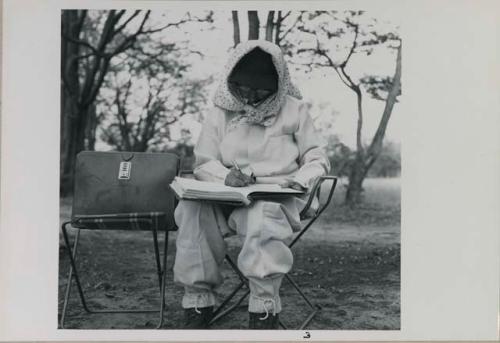 Lorna Marshall wearing clothing and head covering to protect against tsetse flies, sitting and writing in her notebook