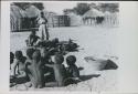 Group of children playing a game and Rose standing, with huts in background

