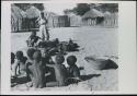 Group of children playing a game and Rose standing, with huts in background

