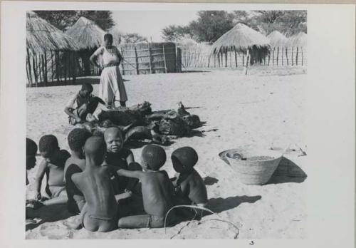 Group of children playing a game and Rose standing, with huts in background

