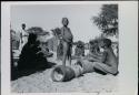 Group of children sitting, and one is playing the le!goma; and two pestles on ground


