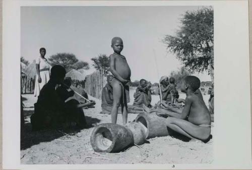 Group of children sitting, and one is playing the le!goma; and two pestles on ground


