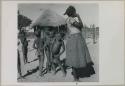 Woman standing, with children behind her and hut in background