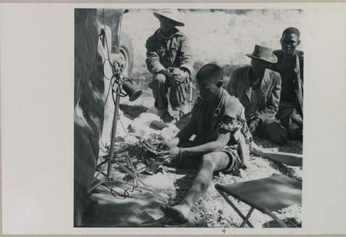 Man playing sanza and being recorded, and Kernel Ledimo and other men sitting near him
