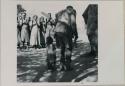 Man dancing with rattles, close-up, child standing next to him, and women clapping in background