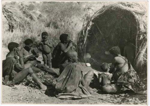 "Living places, shelters": Group of people sitting in front of the skerm of Gau of Band 2, including "Old /Gasa," //Khuga (wife of /Naishi), a woman stringing beads, "Little /Qui," and "Old Gau" from Band 1 (print is a cropped image)