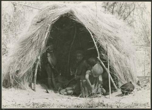 "Living places, shelters": Group of people sitting and a child standing in a skerm, with a water bag made from an animal stomach hanging in the entrance (print is a cropped image)