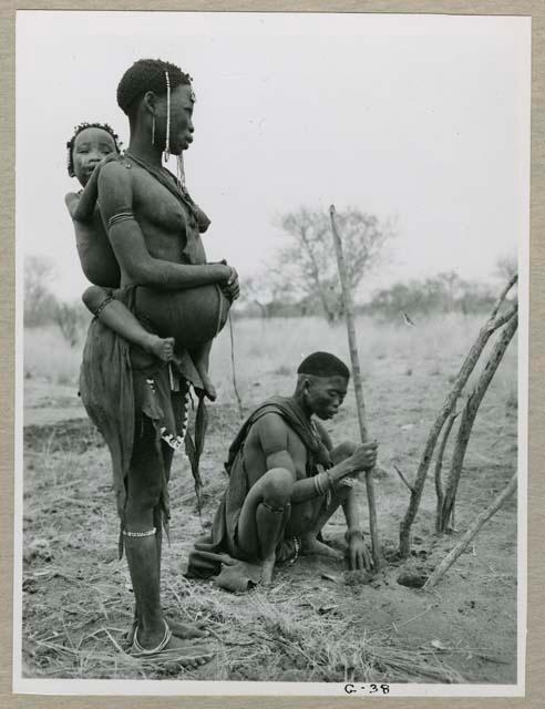 "Living places, shelters": //Kushay (wife of "/Qui Navel") standing, with "Little ≠Gao" holding on to her back, !Ungka sitting on the ground using her digging stick to dig a hole for a branch of a skerm frame (print is a cropped image)