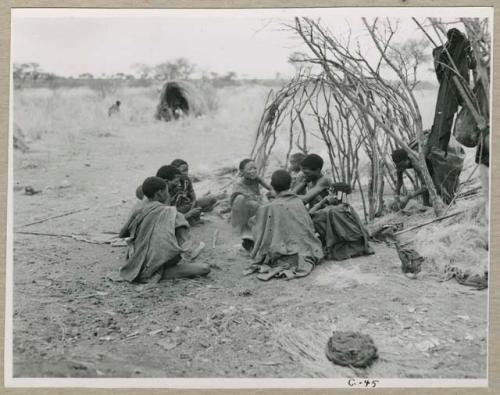 "Living places, shelters": "Gao Medicine" sitting in front of a partially constructed skerm with his family members, including /Naoka (his first wife) and ≠Toma, Di!ai is working on the skerm behind them (print is a cropped image)