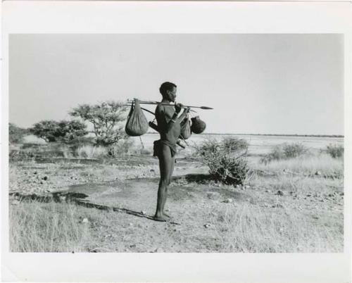 [No folder title]: /Ti!kay carrying bags on a carrying stick and an assegai, with Gautscha Pan in the background (print is a cropped image)