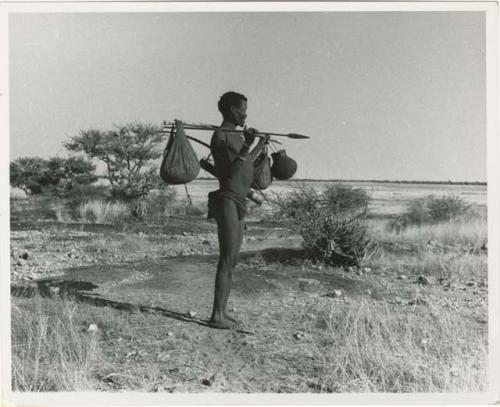 [No folder title]: /Ti!kay carrying bags on a carrying stick and an assegai, with Gautscha Pan in the background (print is a cropped image)