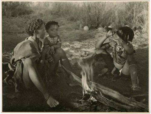 [No folder title]: Woman sitting with a child and a girl by a fire (print is a cropped image)