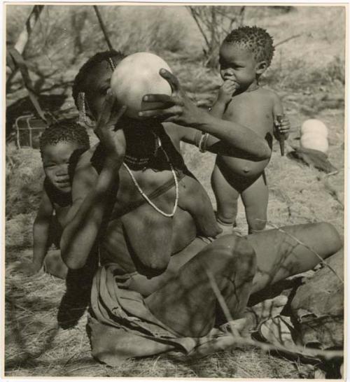 "Children in groups": /Naoka, wife of Ti!kay, drinking from an ostrich eggshell, with !Nani behind her, and /Khoa, her daughter, standing beside her (print is a cropped image)