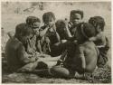 "Children in groups": Group of boys and girls sitting close together, boys are making toy cars (print is a cropped image)