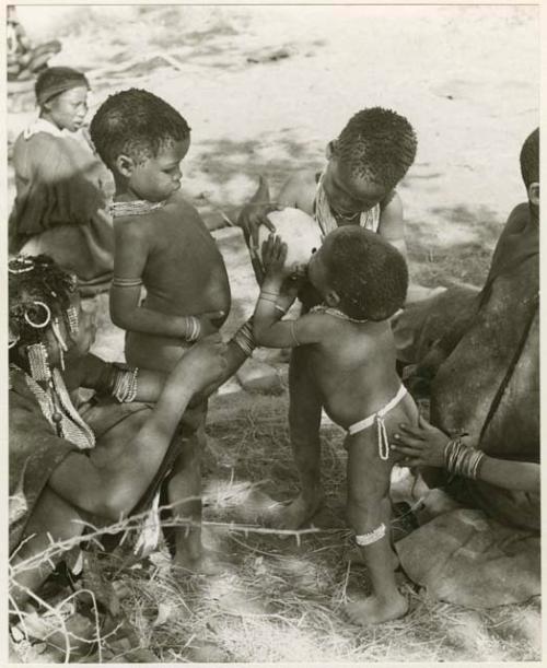 [No folder title]: /Gaishay and an unidentified, young woman helping Debe drink from an ostrich eggshell while Bau watches and another hand steadies Debe; other unidentified people in the background (print is a cropped image)