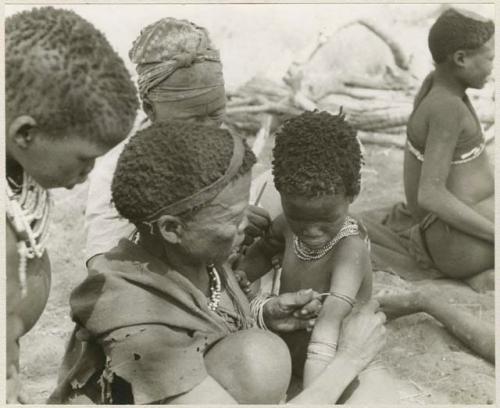 [No folder title]: Group of women and children including !Ungka and an unidentified woman removing ornaments from /Khoa (/Qui's daughter) (print is a cropped image)