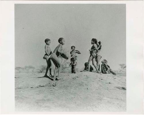 "Boys and girls playing together": Boys and girls dancing on a ledge by a waterhole, one boy playing a //guashi