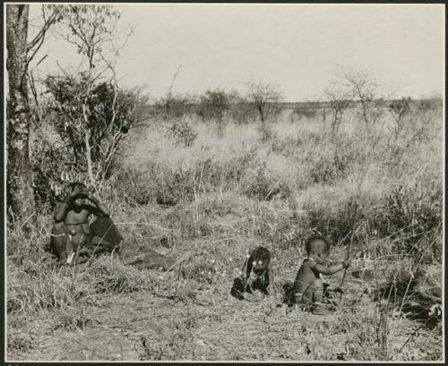 "Children playing gathering": Two women sitting in grass and two children playing gathering veldkos, one holding a digging stick (print is a cropped image)