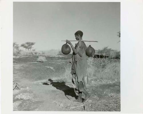 "Water": Woman carrying two water bags made from animal stomachs over a digging stick on her shoulder (print is a cropped image)