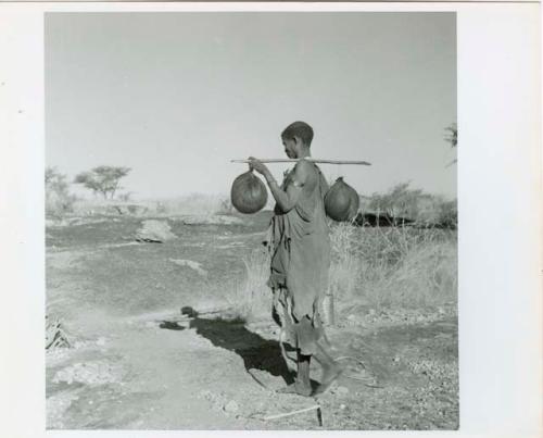 "Water": Woman carrying two water bags made from animal stomachs over a digging stick on her shoulder (print is a cropped image)