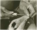 Person sewing, close-up of hands using awl to make a hole for putting a cord through (print is a cropped image)