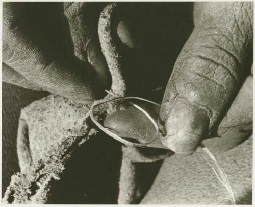 Person sewing, close-up of hands putting unrolled vegetable fiber through a hole in leather (print is a cropped image)