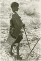 DaSi!Na in profile standing and leaning against digging stick, showing apron around her waist (print is a cropped image)