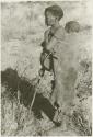 Tsekue in a kaross with N!whakwe tied to her back, standing holding a digging stick (print is a cropped image)