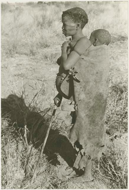 Tsekue in a kaross with N!whakwe tied to her back, standing holding a digging stick (print is a cropped image)