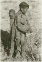 Tsekue wearing a kaross, with N!whakwe tied to her back, holding a digging stick, full figure (print is a cropped image)