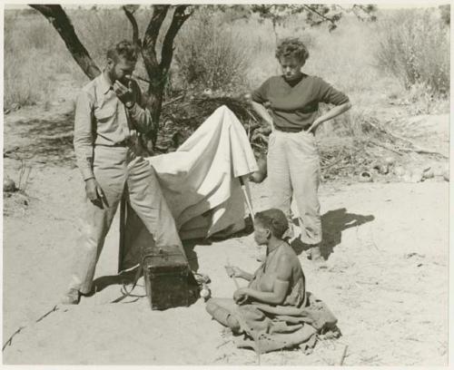 Tsekue making musical sounds and rhythms using stiff grass, with Daniel Blitz and Elizabeth Marshall Thomas preparing to set up synchronized sound recordings (print is a cropped image)