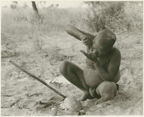 Oukwane drinking, using one hand to squeeze juice into the palm of the other hand, full figure (print is a cropped image)