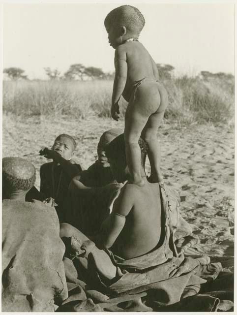 N!whakwe standing on his mother Tsekue's shoulder, seen from behind (print is a cropped image)