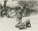 Three boys playing in the sand; women sitting in the background (print is a cropped image)