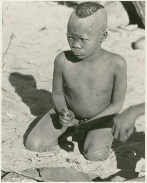 /Gaiamakwe striking a rhythm on the lower string of musical bow with a stick (print is a cropped image)