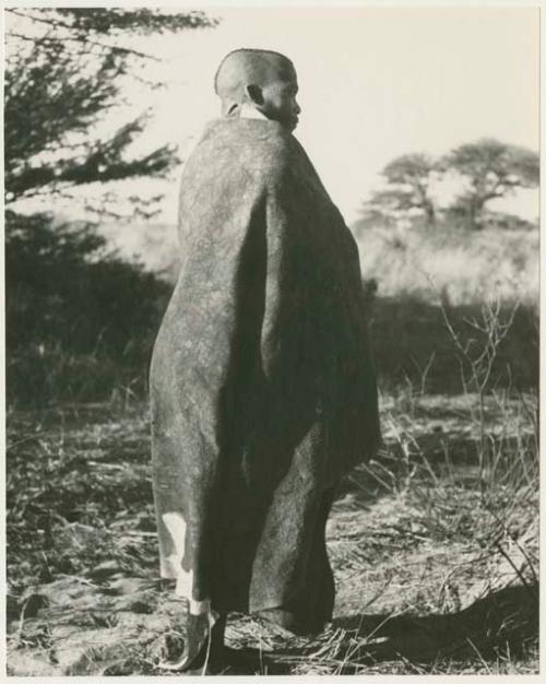 Visiting man wrapped in a big kaross, seen from behind, full figure standing (print is a cropped image)