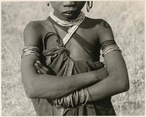 N!ai with her arms crossed, showing necklace, fiber strands on upper arm, and wrist bracelets made of thong, grass, and wire (print is a cropped image)