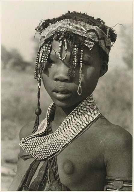 Portrait of N≠isa wearing ostrich eggshell beads around her neck, headband of Western beads on her head, and a long band made of a tsi nut and beads hanging on the right side of her face (print is a cropped image)