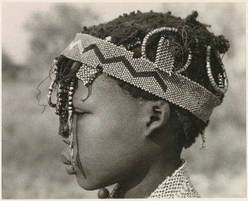 Close-up profile of N≠isa wearing ostrich eggshell beads around her neck, headband of European beads on her head, and a long band made of a tsi nut and beads hanging on the right side of her face (print is a cropped image)