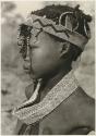 Profile portrait of N≠isa wearing ostrich eggshell beads around her neck, headband of European beads on her head, and a long band made of a tsi nut and beads hanging on the right side of her face (print is a cropped image)