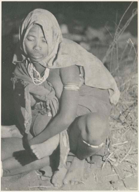 //Kushay squatting with a kerchief around her head, seen from the front (print is a cropped image)