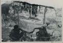 ǂToma ʒo and //Kushay sitting by a fire, beneath a trestle with meat hanging from it

